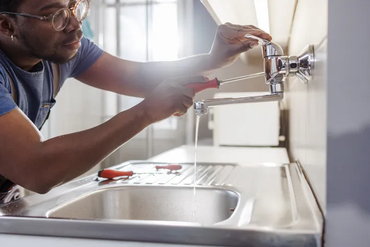 A plumber uses a screwdriver to repair a chrome kitchen faucet over a stainless steel sink.