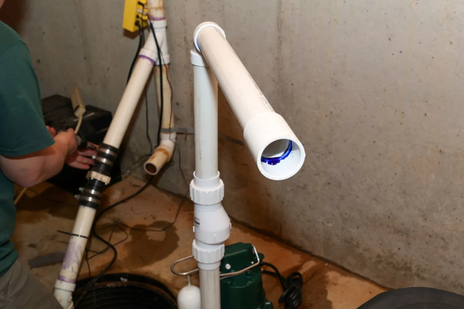 A white PVC discharge pipe of a sump pump is visible in the foreground while a person performs a sump pump repair in the background.