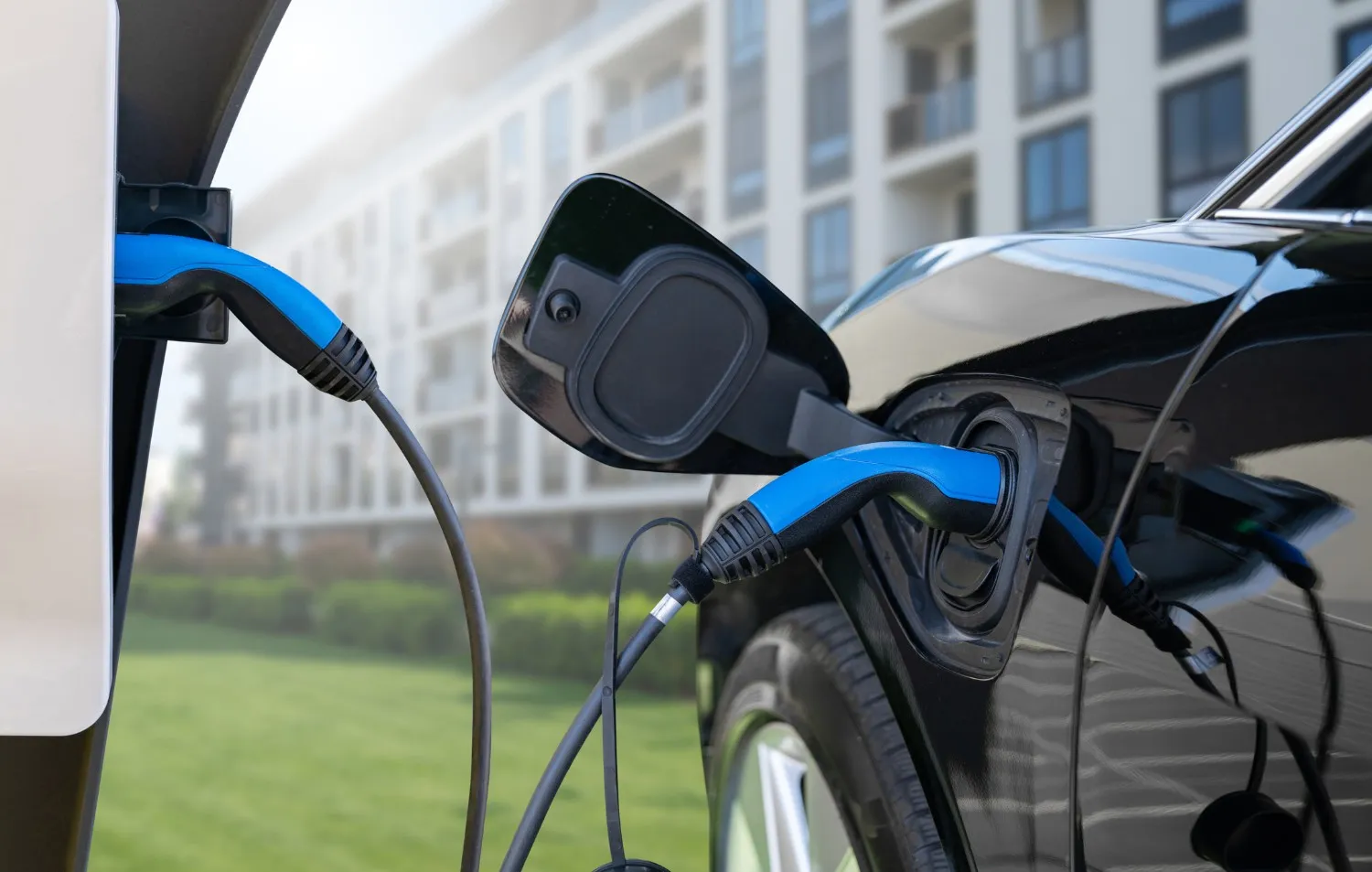 A black electric vehicle is connected to an EV charging station with a blue cable in front of a modern apartment building.