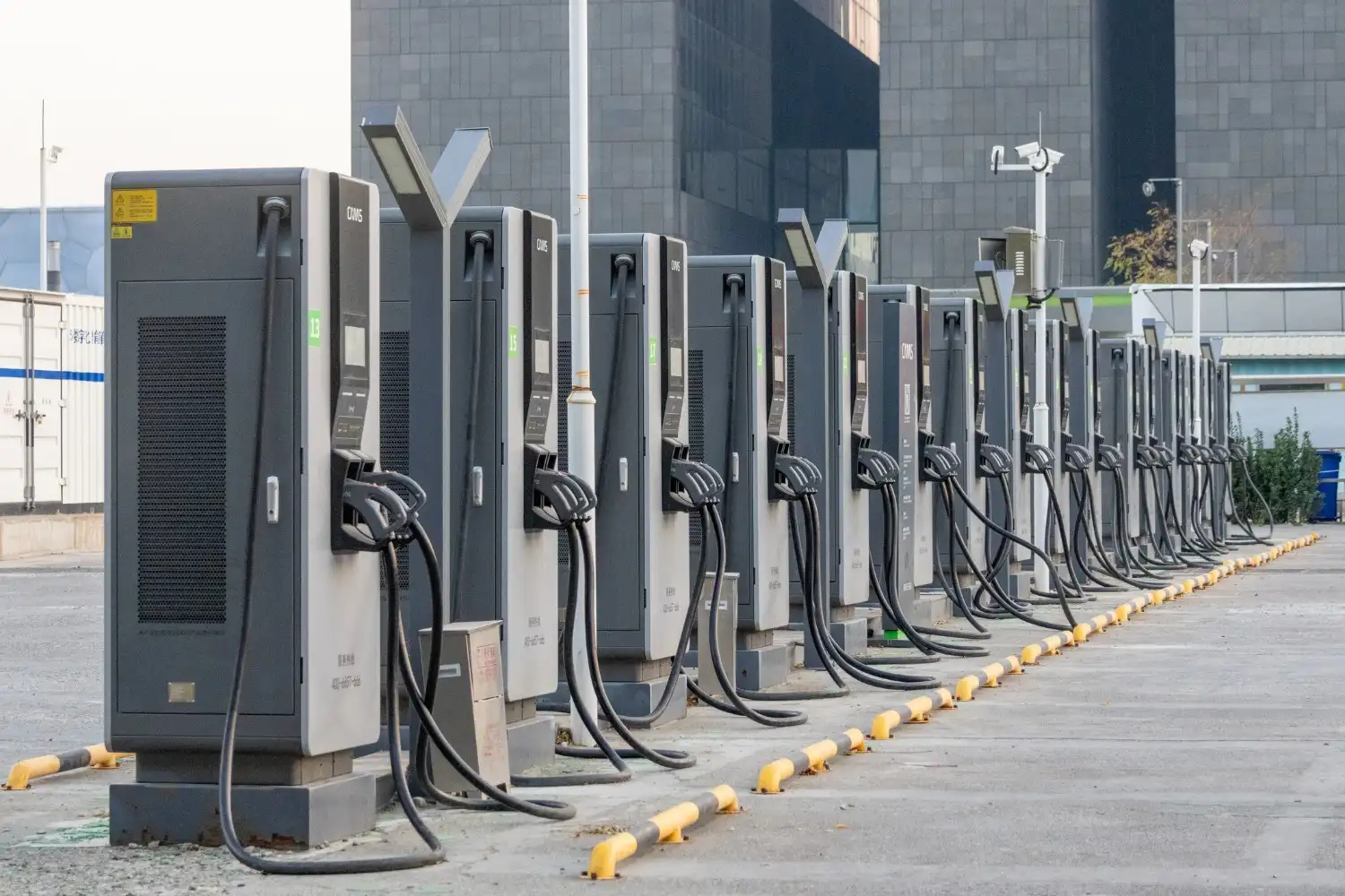 A long row of unoccupied EV charging stations with attached cables lines an outdoor parking lot in front of tall buildings.