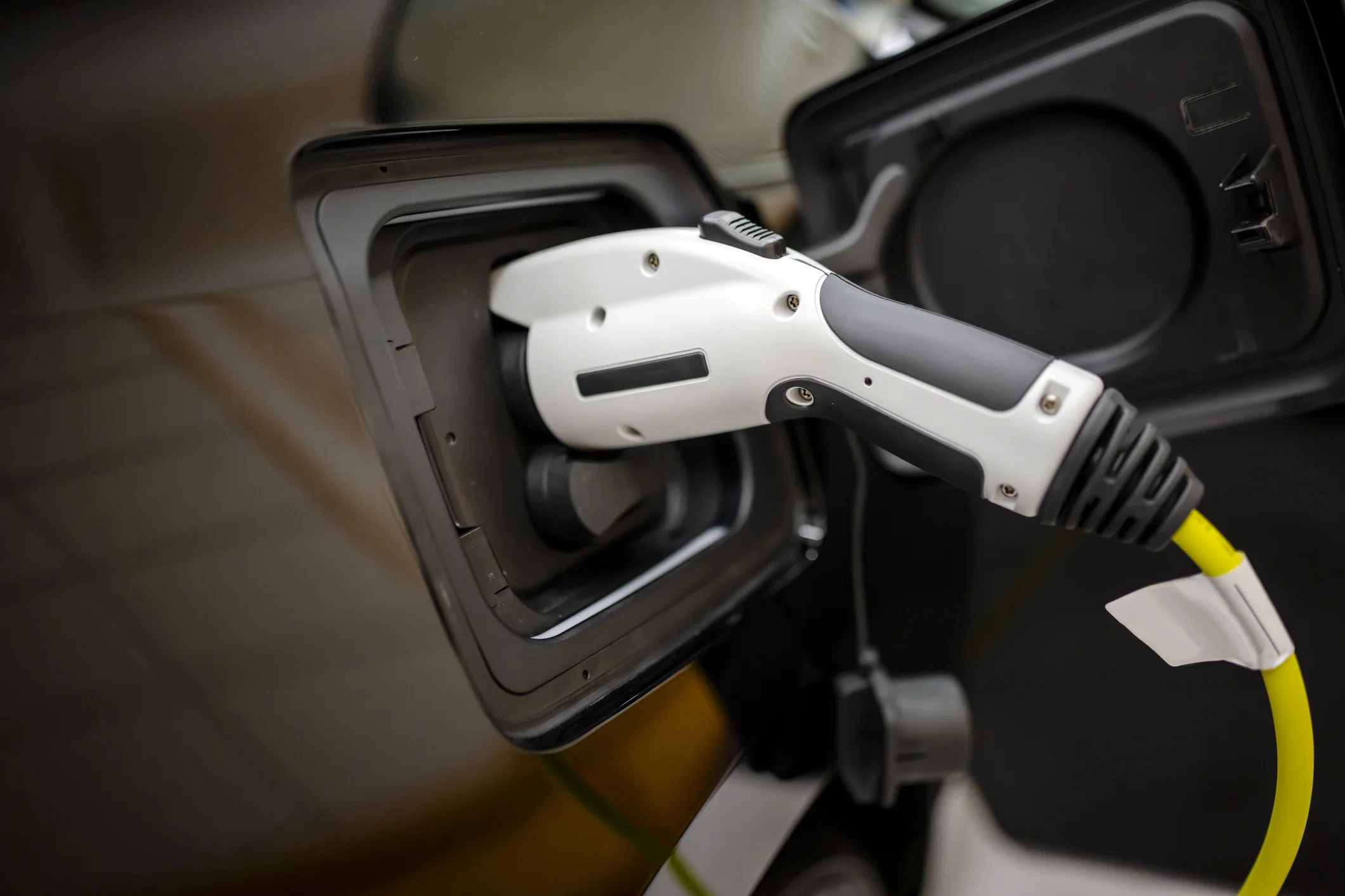  A close-up view of a white and black electric vehicle (EV) charging plug inserted into the charging port of a dark-colored car. The port is open, revealing the connection. A bright yellow cable is attached to the plug.