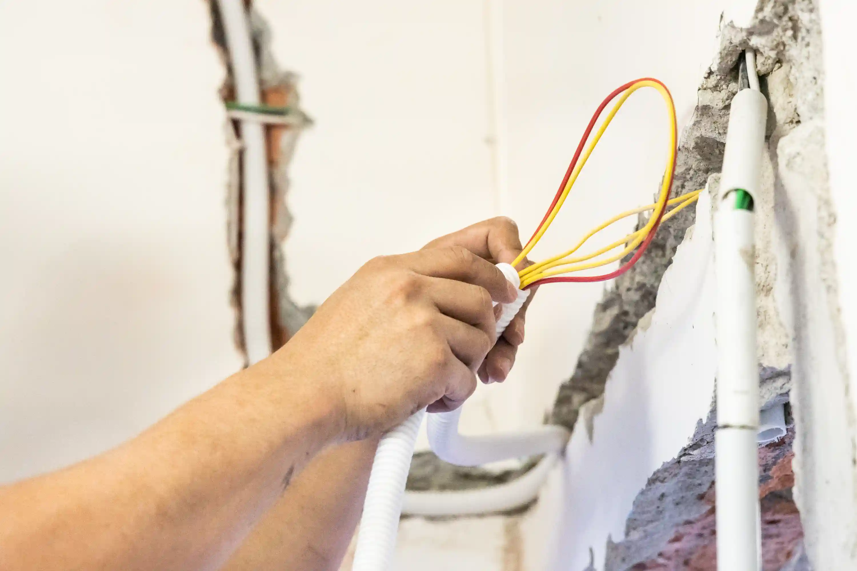  A close-up shot of a worker's hands pulling red and yellow electrical wires through a white flexible corrugated conduit. The wires are being fed through a section of wall that has been broken open, revealing the original brickwork and plaster, indicating a house rewiring or renovation project.