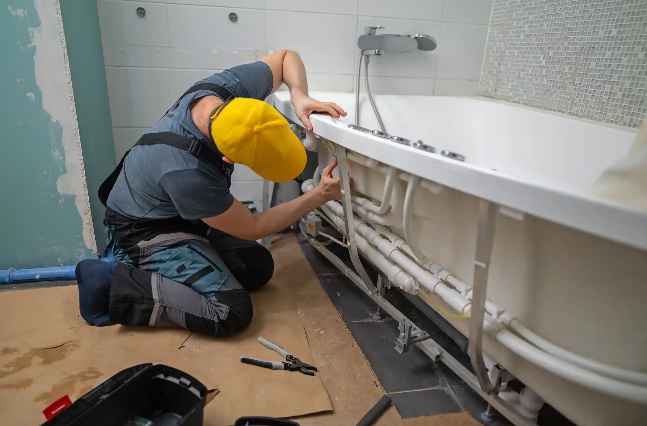 A plumber wearing a yellow hard hat and work clothes is kneeling on the floor, working on the exposed piping and wiring underneath a white bathtub during a bathroom renovation. The floor is covered with a protective layer of cardboard, and tools are visible in the foreground.