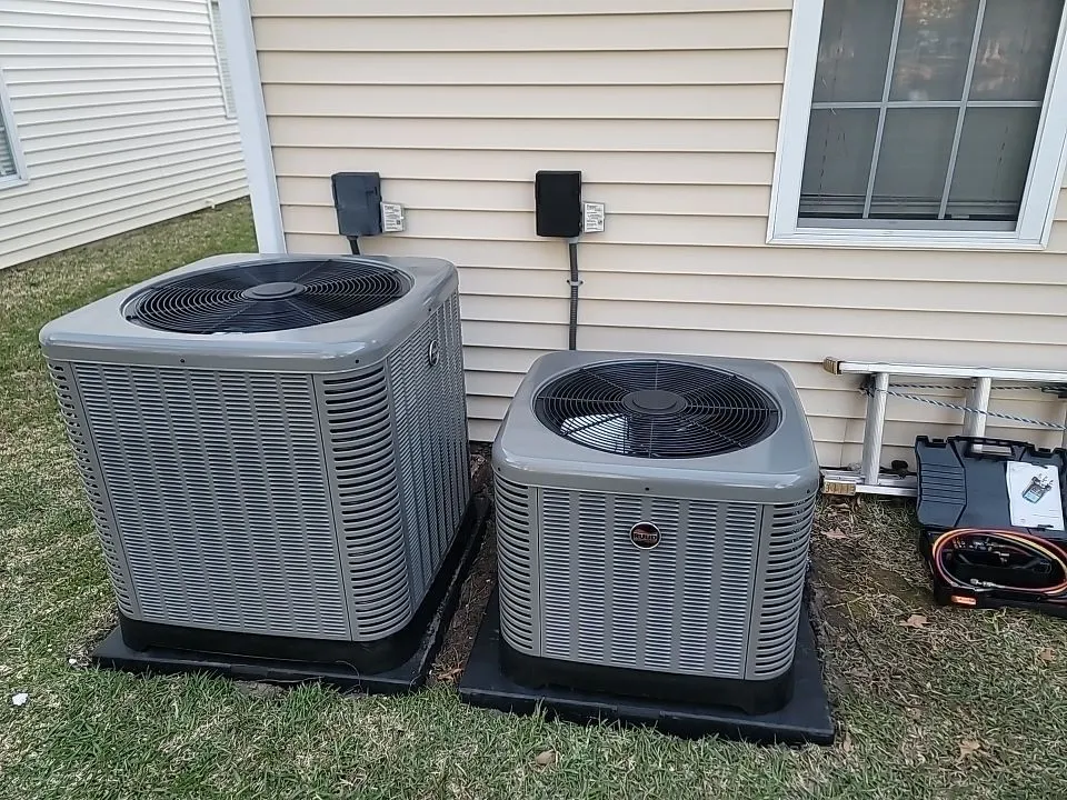 Two gray outdoor air conditioning units sit on concrete pads beside a beige house.