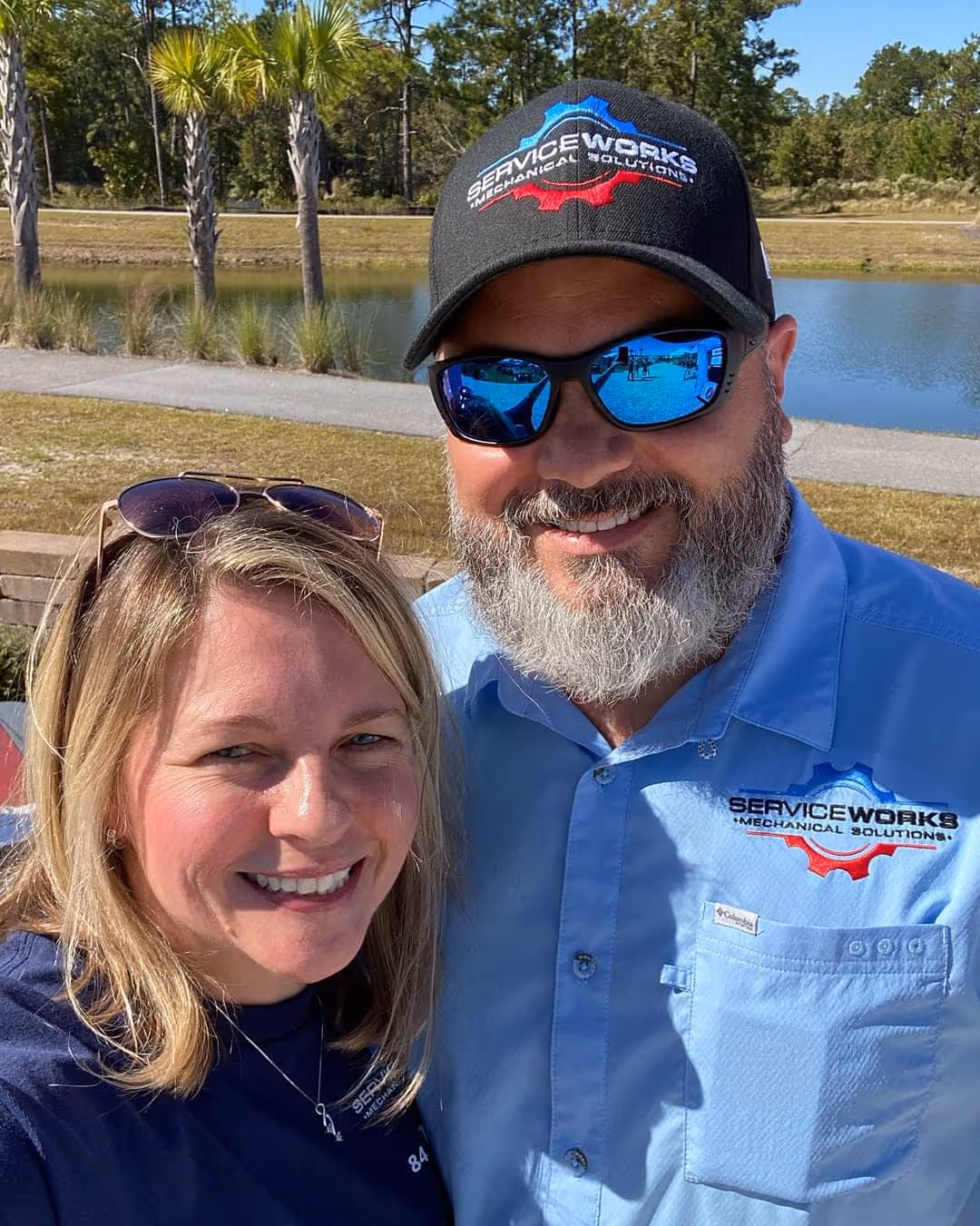 A smiling man and woman outdoors on a sunny day. The man wears sunglasses, a cap, and a blue shirt with a company logo.
