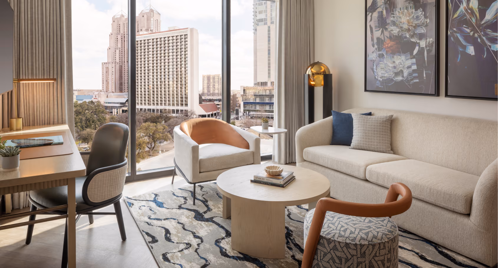Corner suite living area with curved sofa, accent chairs, round coffee table, and large window showing the San Antonio skyline