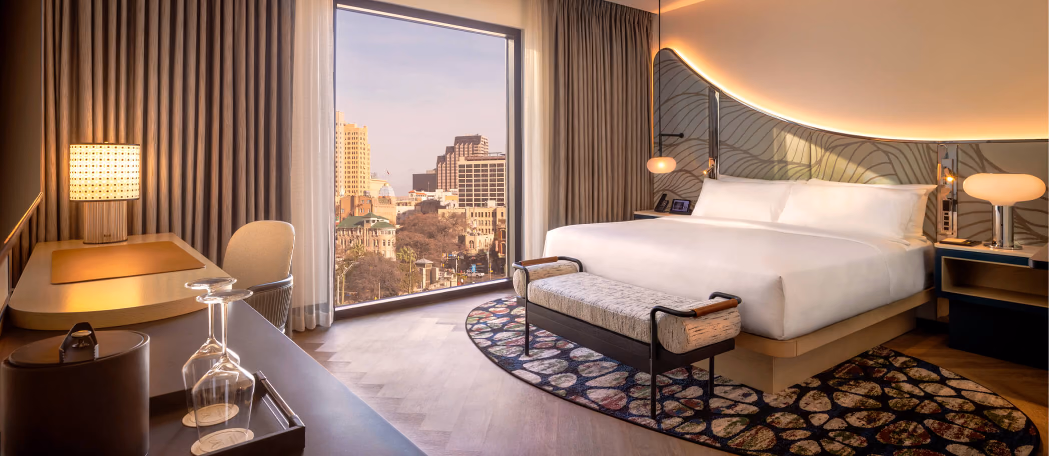 King hotel room with bed, desk, woven lamp, and floor-to-ceiling window overlooking downtown San Antonio at sunset