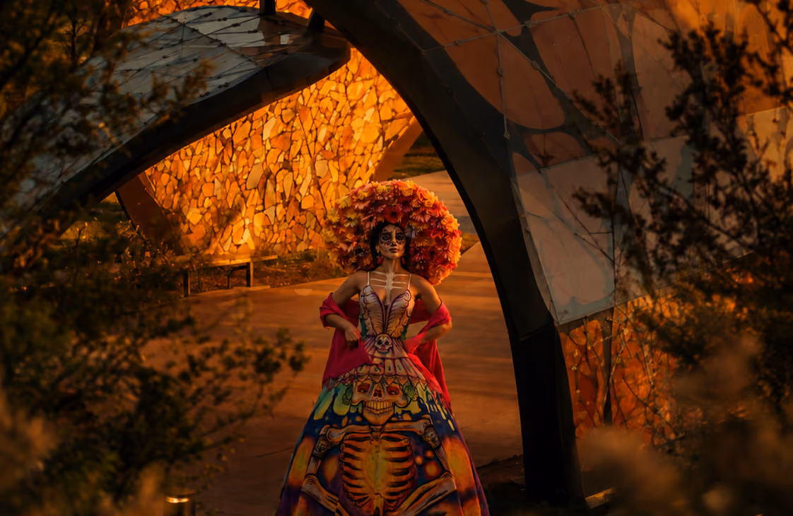 Woman in colorful Day of the Dead dress and large flower crown stands under a glowing arch.