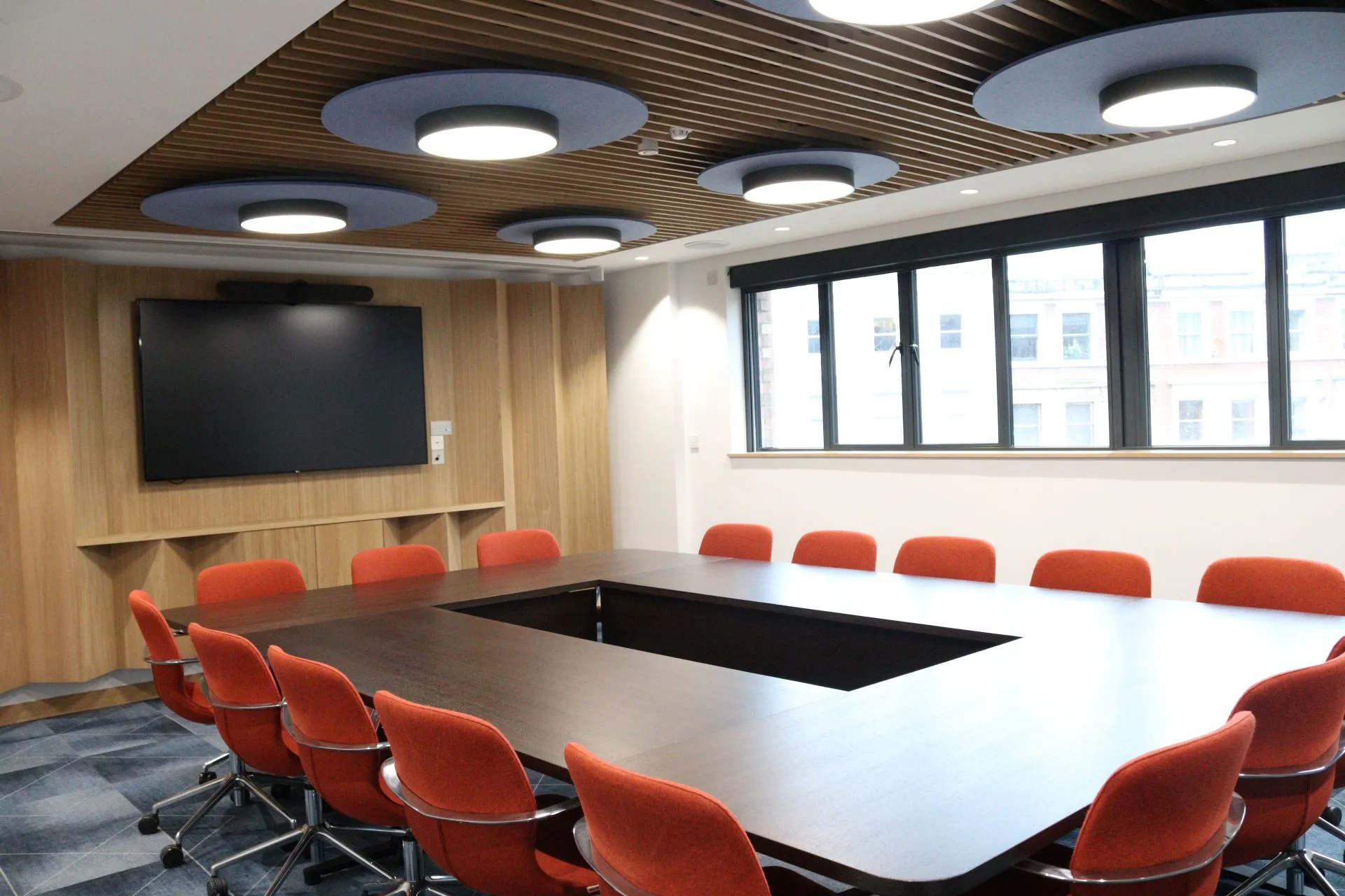 One of the meeting spaces within the Henley Suite. A large meeting table with colourful chairs, wood panelling on the walls and a TV on the wall. A large window brings light into the room.