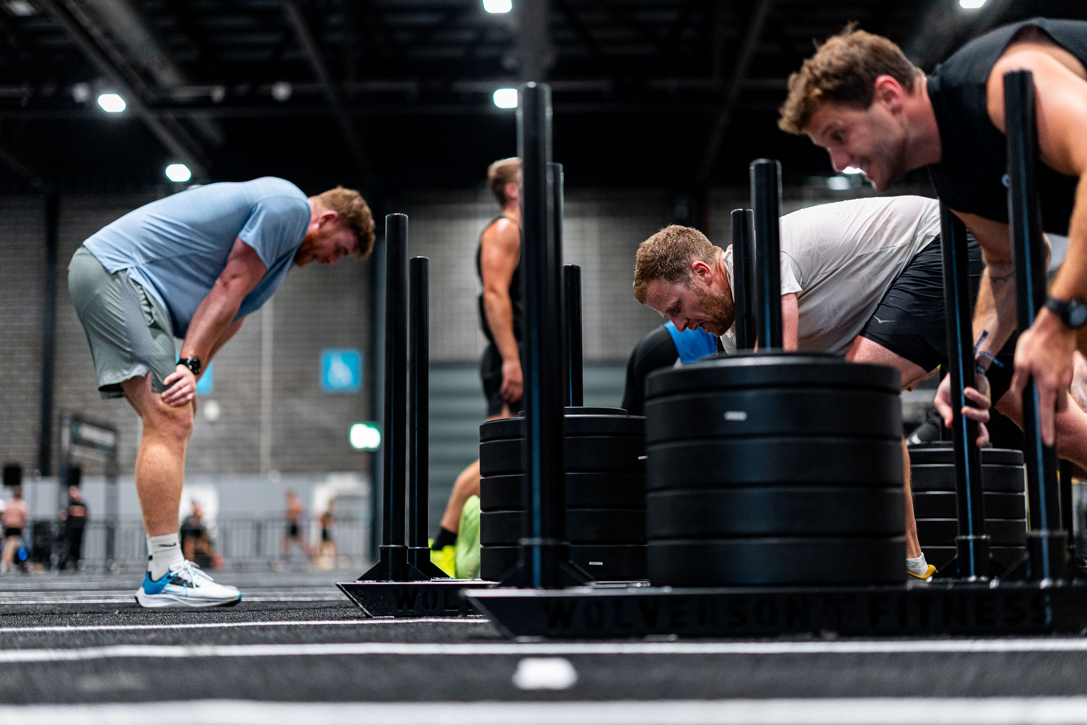 Athletes compete in a fitness challenge, pushing heavy weighted sleds across the floor, while one participant in a blue shirt pauses to catch his breath.