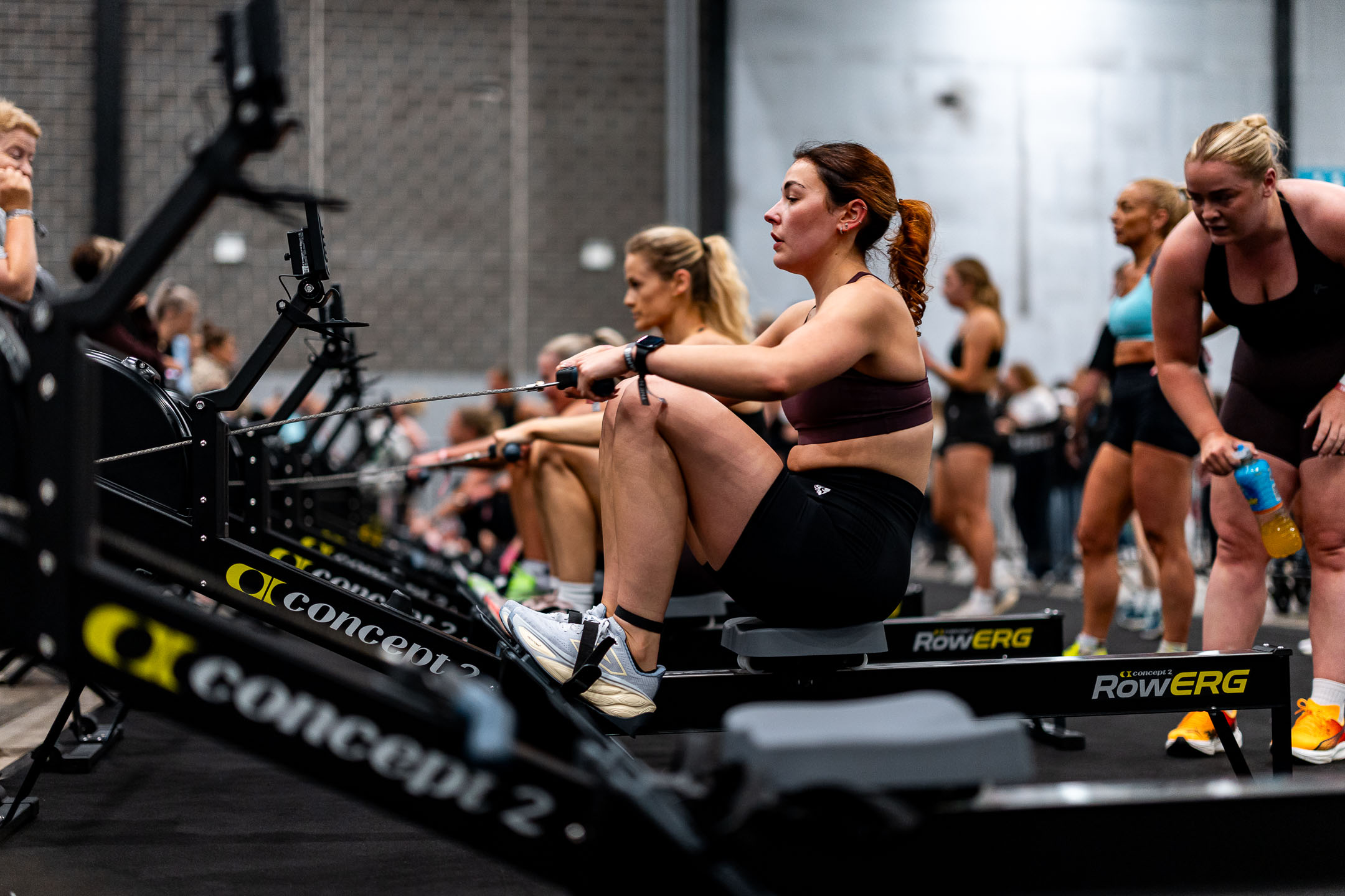 A woman in athletic wear rows intensely on a Concept2 rowing machine during a fitness competition, with other athletes competing alongside her and spectators in the background.