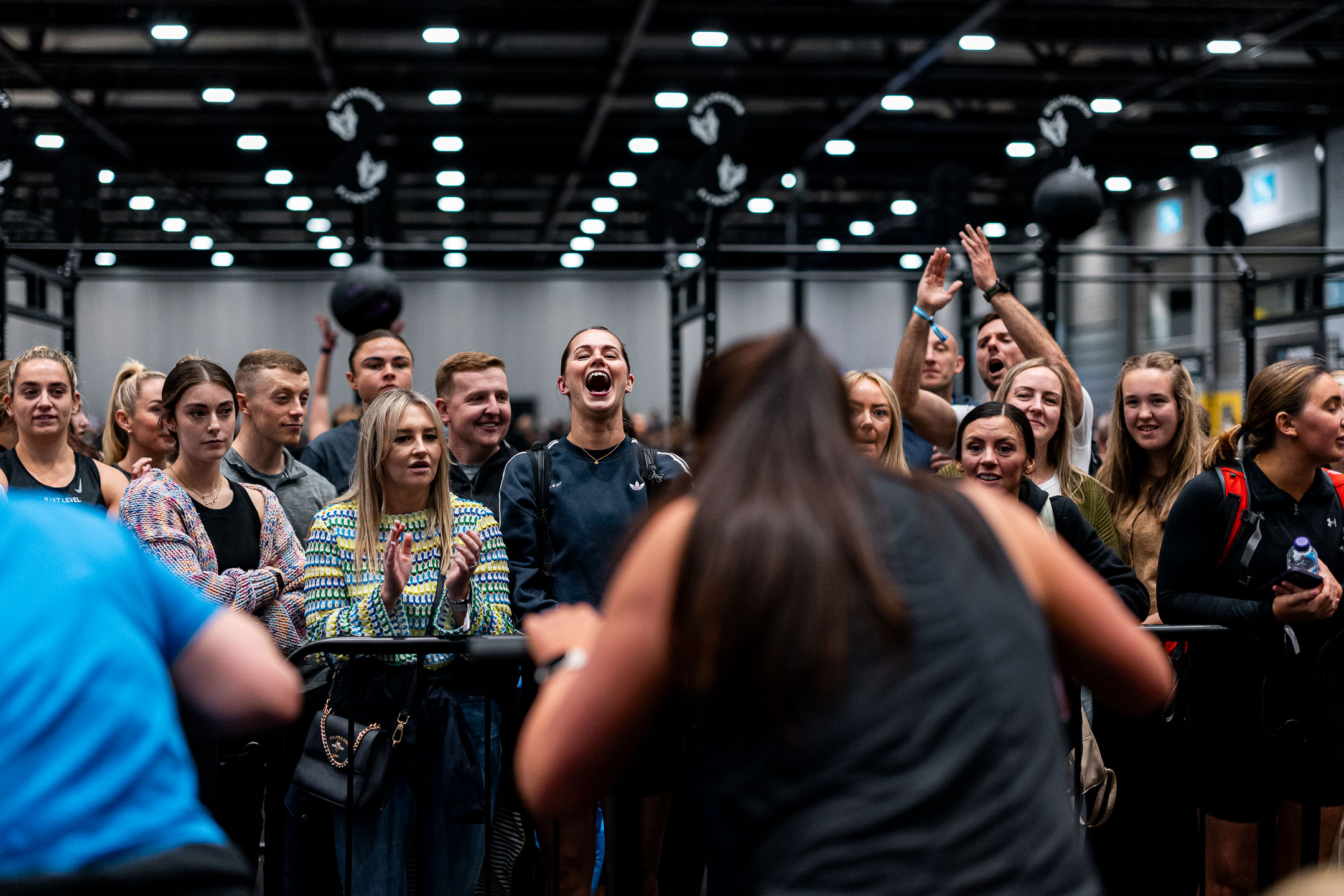 A lively crowd of spectators cheers and claps behind a barrier at a fitness event, encouraging athletes as they compete on the floor.
