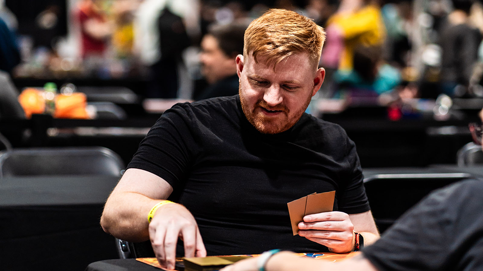 A man with red hair smiles while playing a trading card game at a convention table.