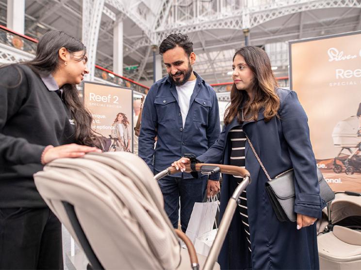 A sales assistant shows a beige stroller to a couple at a baby products exhibition, with branded displays in the background.