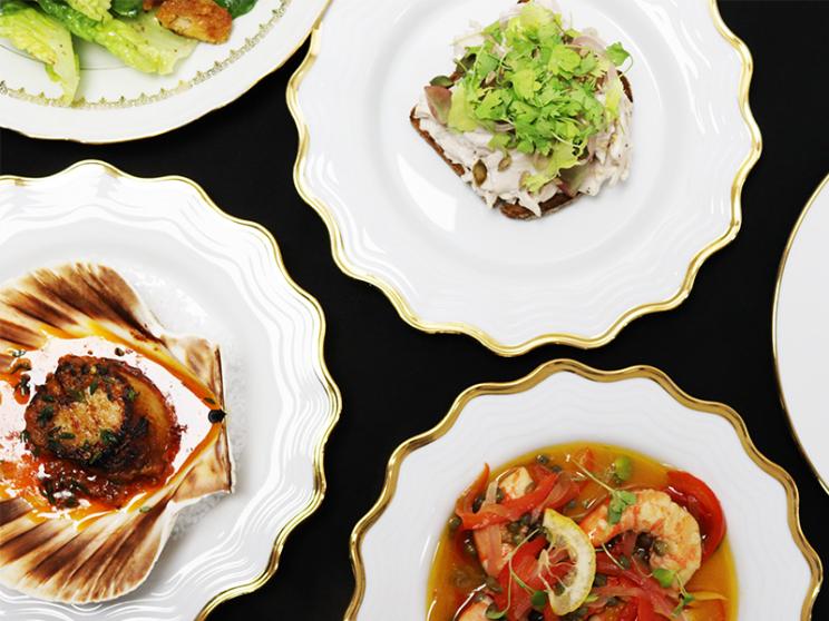 A selection of gourmet dishes presented on elegant white plates with gold trim, including seafood, salad, and small plated starters, displayed against a black tablecloth.