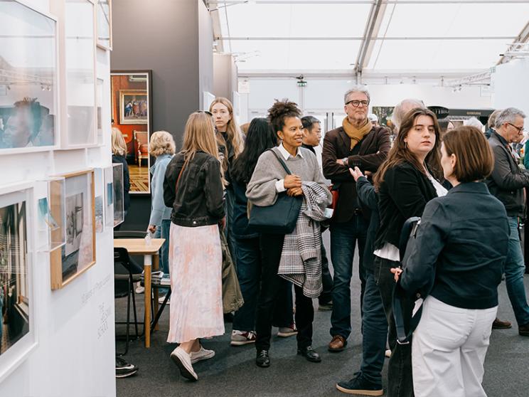 A lively crowd of visitors engages in conversation and browses framed photography at a busy art fair inside a well-lit marquee space.
