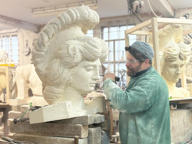 A sculptor wearing safety glasses and a green work jacket carefully carves a large classical-style head sculpture in a workshop, surrounded by tools, stone blocks, and other sculpted pieces.