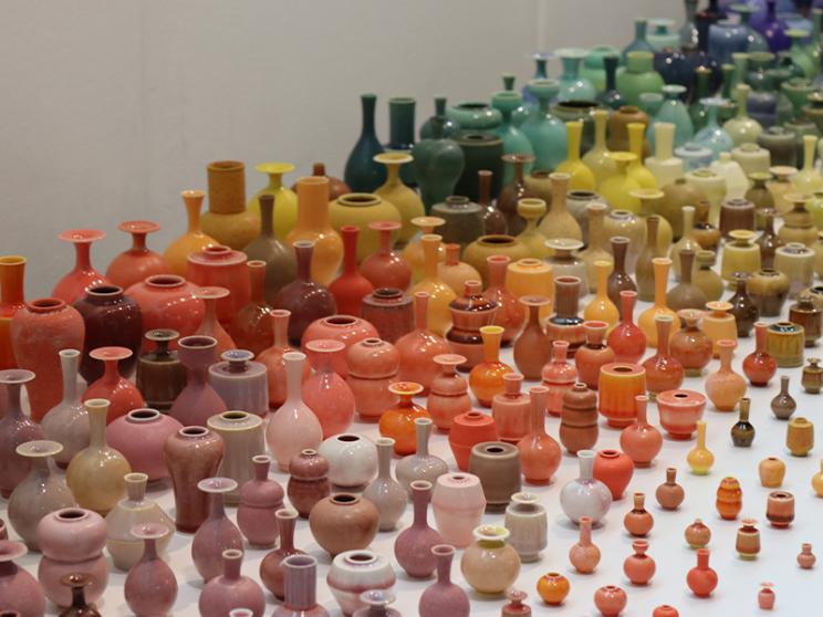 A large display of miniature ceramic vases arranged in a gradient of colors, transitioning from warm tones like reds, pinks, and oranges to cooler hues such as greens, blues, and purples.