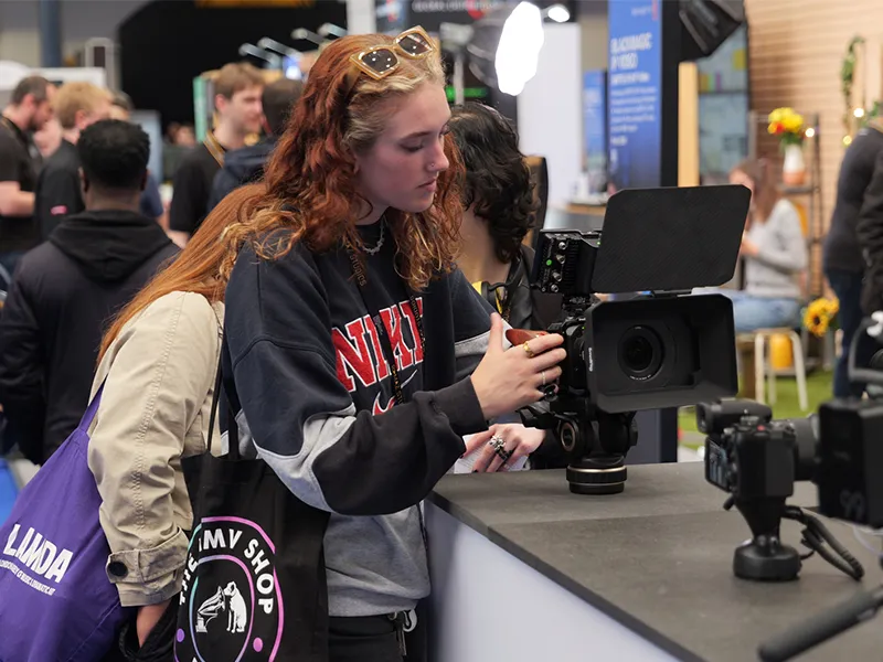 A young woman with red hair and sunglasses on her head looking at a professional video camera on a counter, surrounded by people at MPTS.