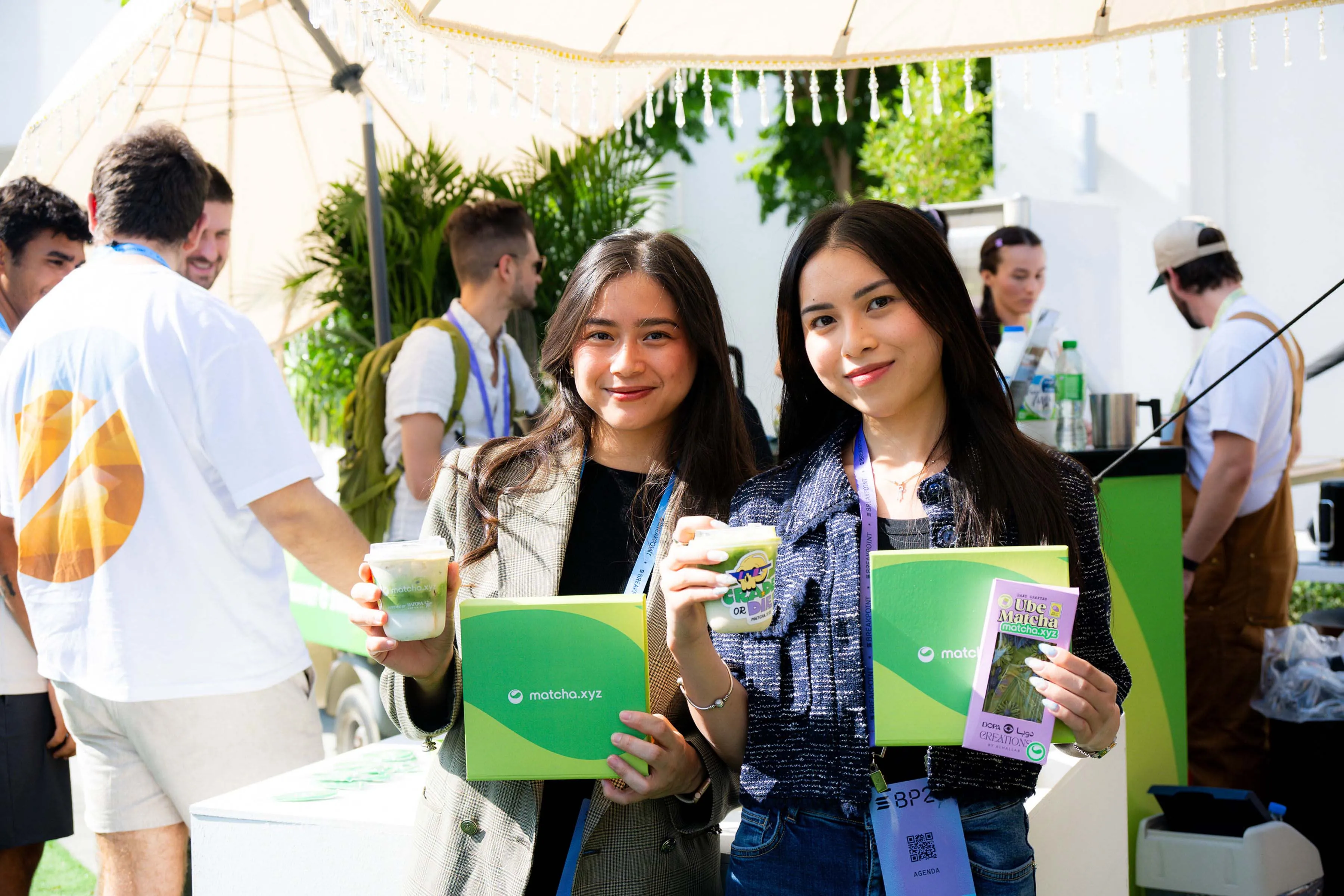 Two young women smiling and holding green boxes and cups at an event with people in the background.