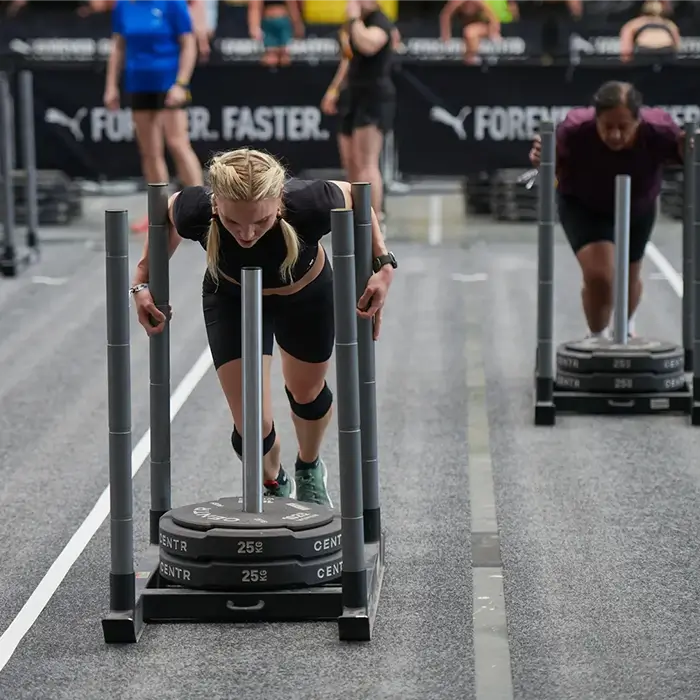 Two women pushing weighted sleds on lanes at HYROX fitness competition, with a blurred background showing other participants and a banner with the words 'Forever. Faster.'