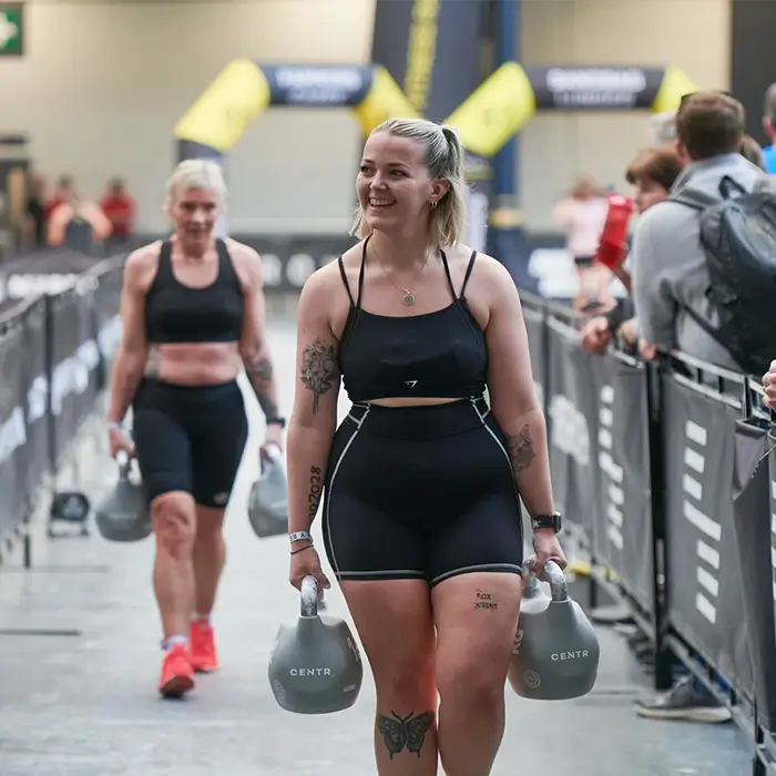 Two women in black athletic wear walking down a gym lane carrying kettlebells, with spectators watching behind barriers at HYROX.