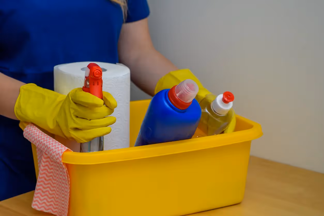 A person holds a yellow bucket filled with cleaning supplies including rags and spray bottles