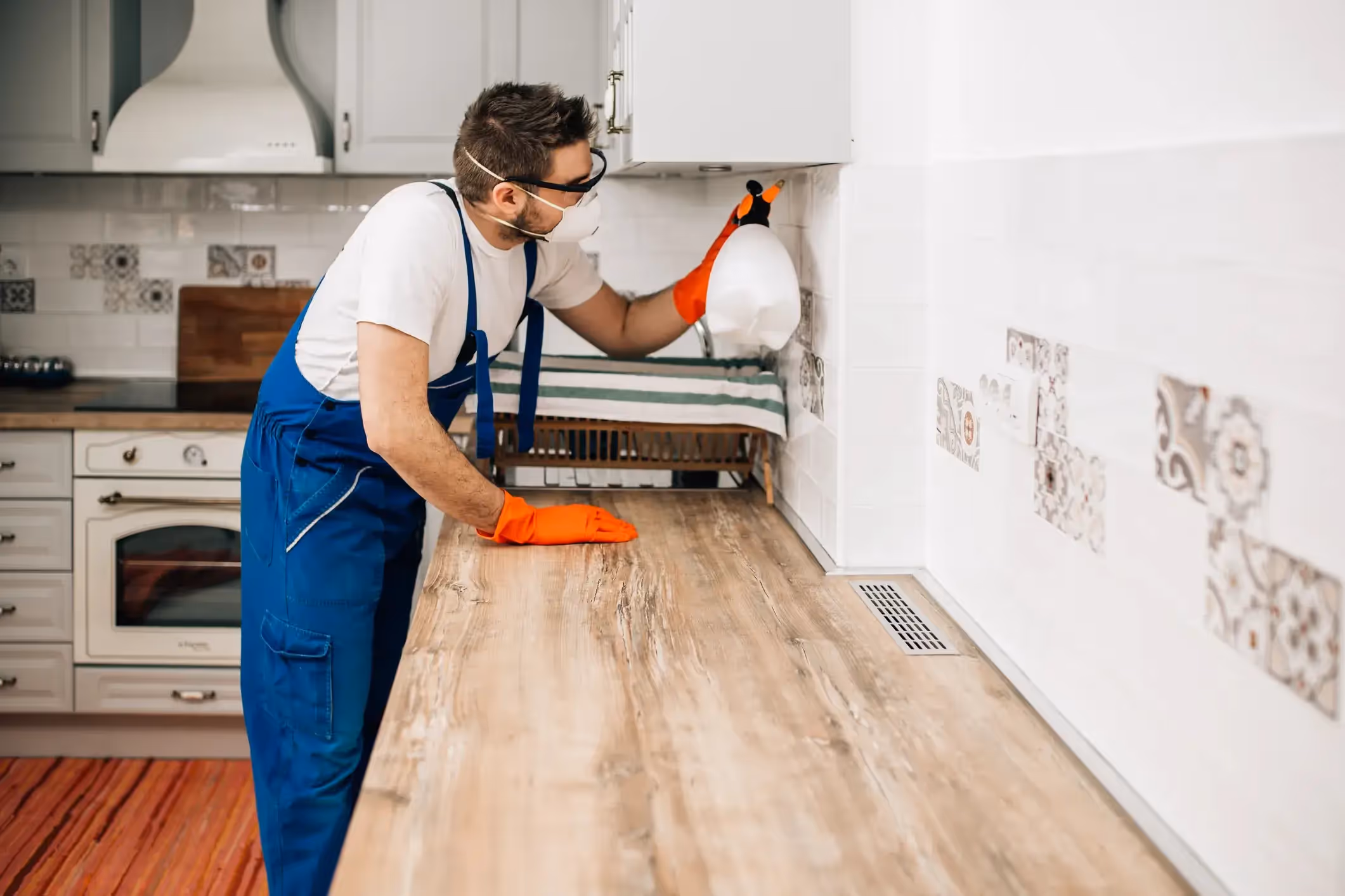 A pest control technician in a blue uniform, mask, and orange gloves sprays under a kitchen cabinet, treating for pests on a wooden countertop.