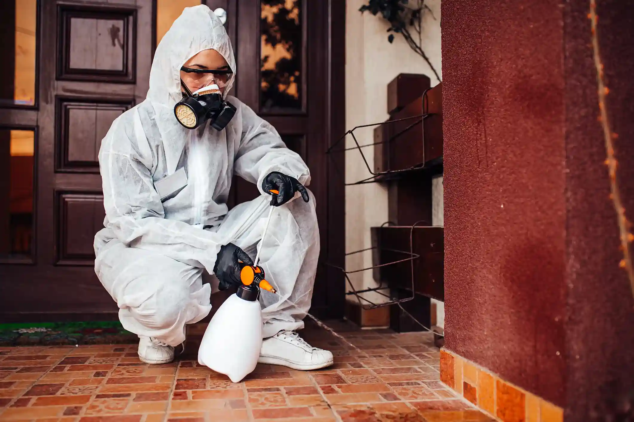 A person in a white protective suit and gas mask crouches to spray a small pump bottle of pesticide near the base of an outdoor wall.