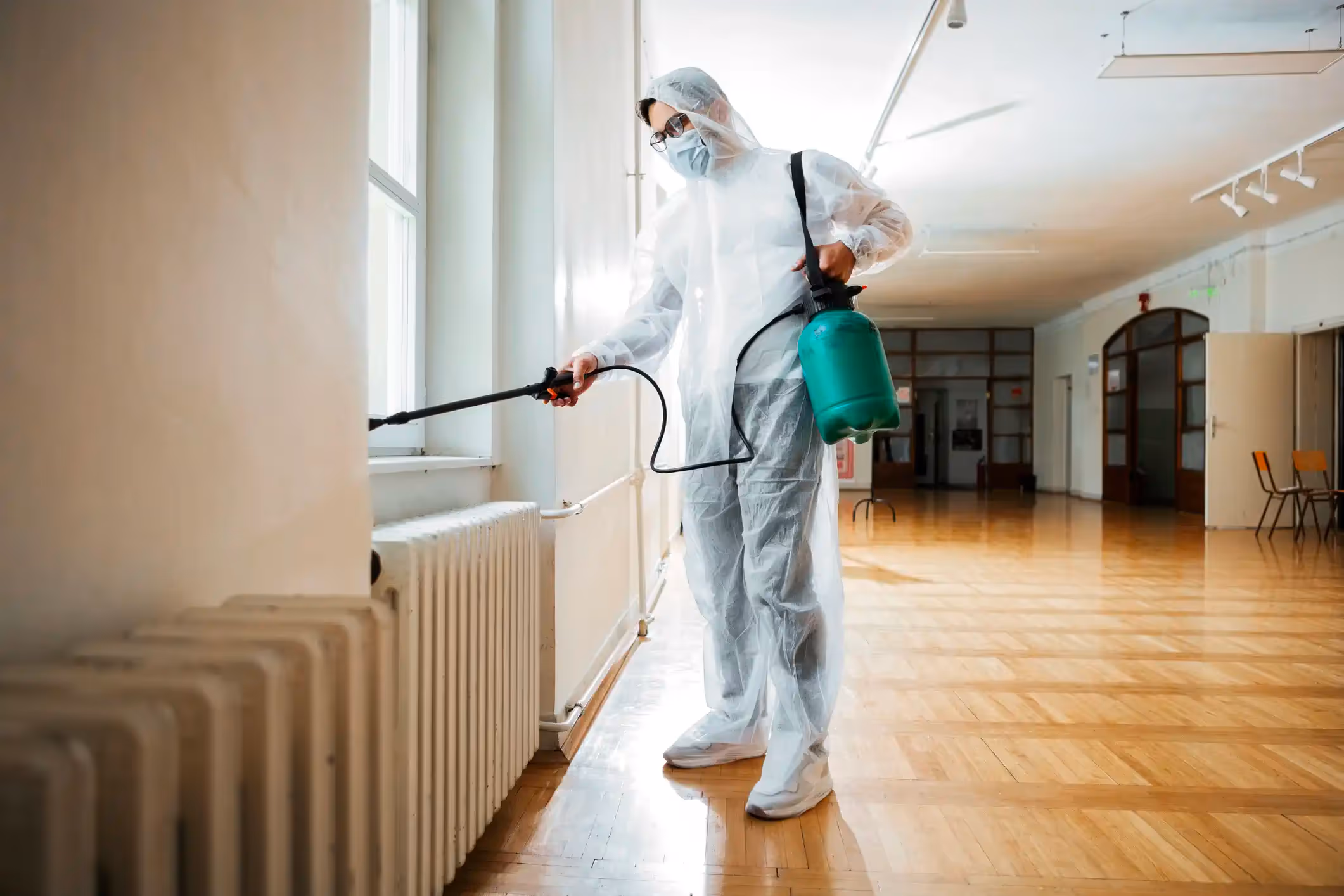 A pest control technician, wearing a white protective suit, face mask, and goggles, sprays along a wall next to a large white radiator. The technician is holding a black spray nozzle and has a bright green tank slung over their shoulder with a black strap. The setting appears to be a school hallway or office building, with a window and wooden floors.
