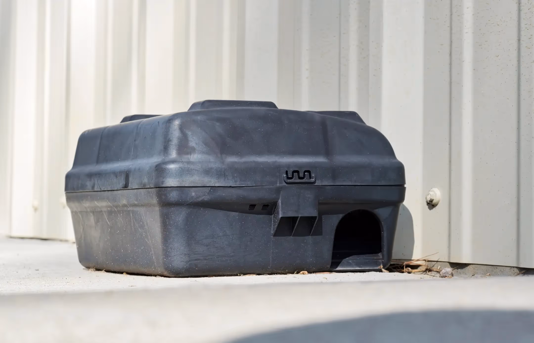 A black plastic bait station for rodent control is sitting on a concrete sidewalk, placed flush against a light-colored, corrugated metal wall. The station has a small, arch-shaped entrance hole on the bottom right. The design suggests it is meant to be tamper-resistant.
