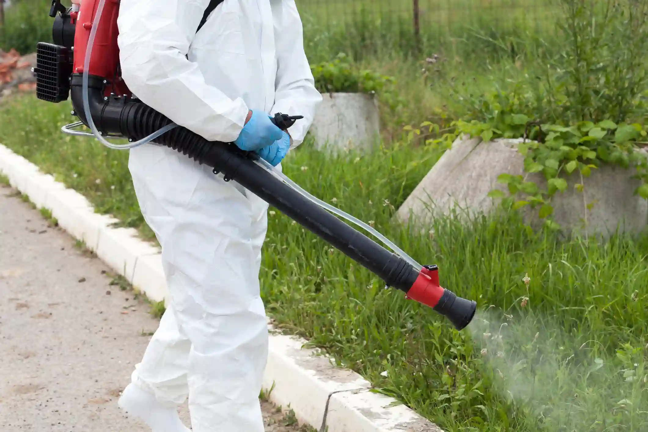 A pest control technician wearing a white protective suit and blue gloves is walking along a grassy area, using a red and black backpack sprayer with a long nozzle to spray a pesticide. The technician's face is not visible.