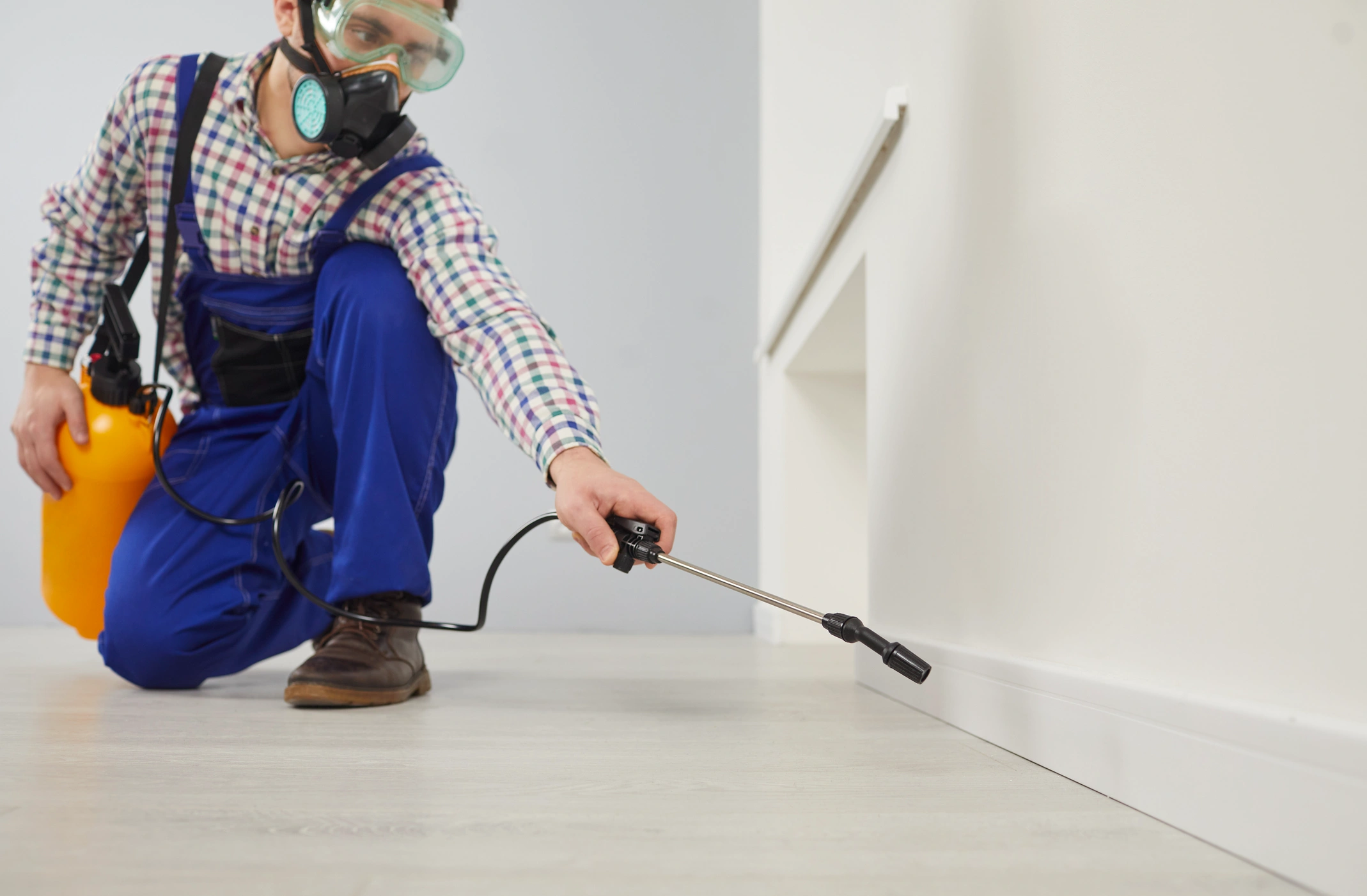 Person in protective gear, including mask and goggles, spraying pesticide along a white baseboard indoors.