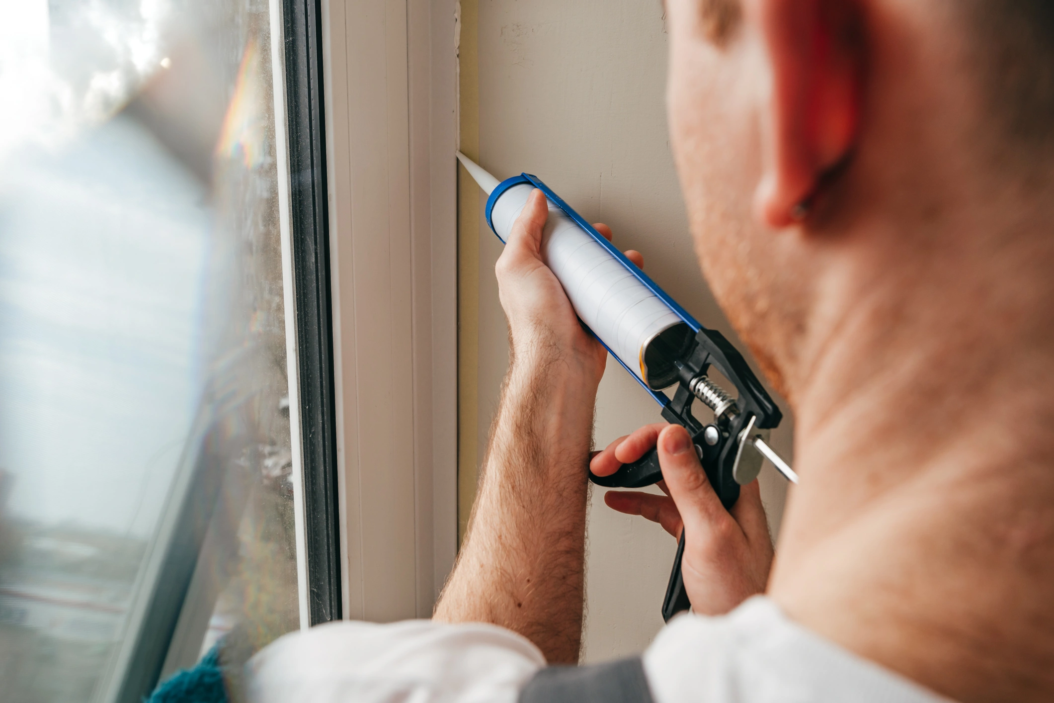 A person applies sealant with a caulking gun around a window frame.