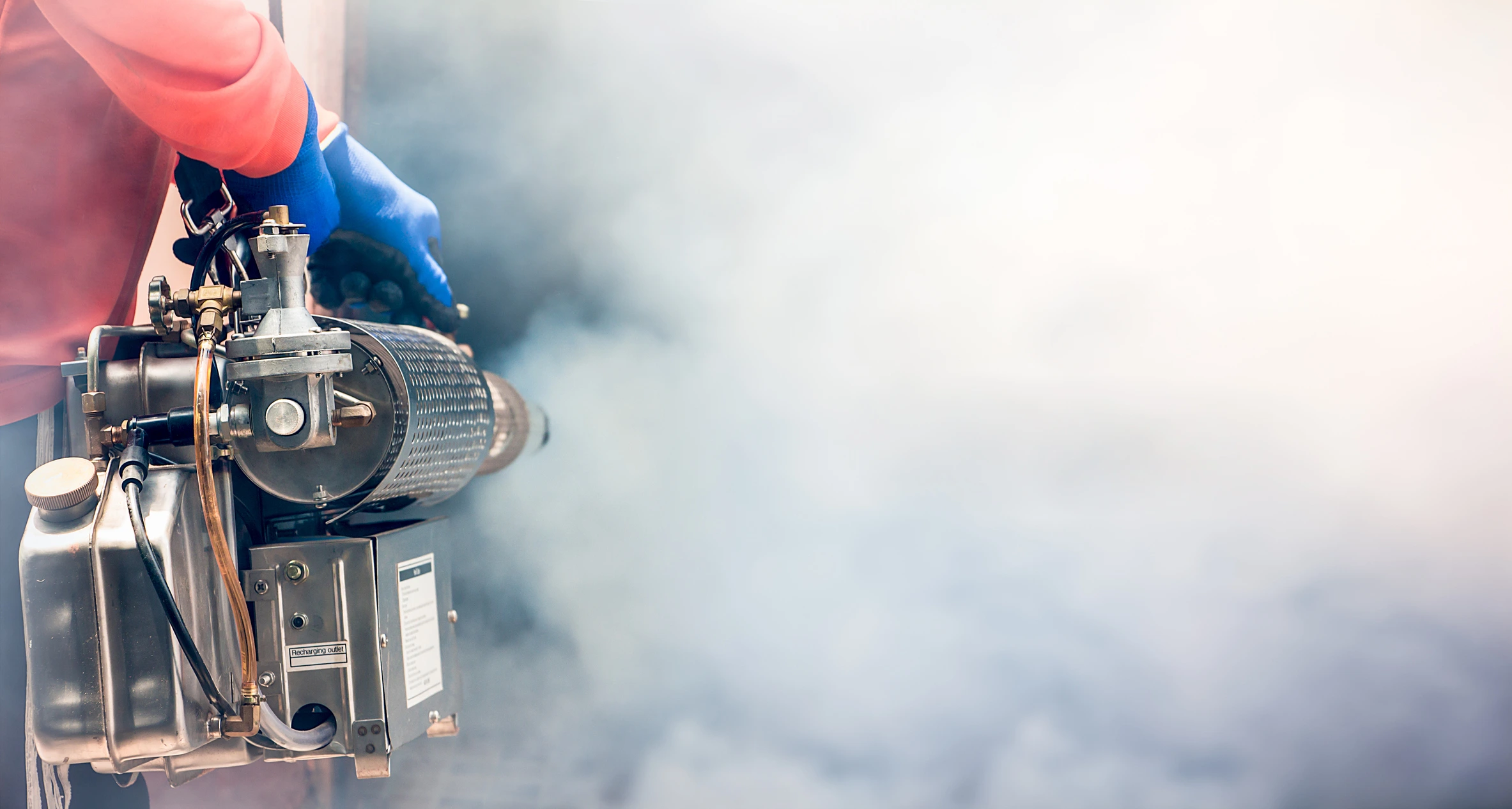 Person in a red shirt and blue gloves using a fogging machine, releasing a thick cloud of mist.
