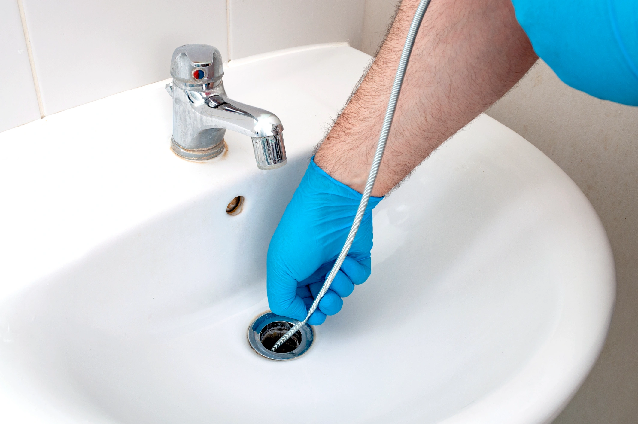 A person wearing blue gloves uses a plumber's snake to unclog a white sink drain. A chrome faucet is visible.