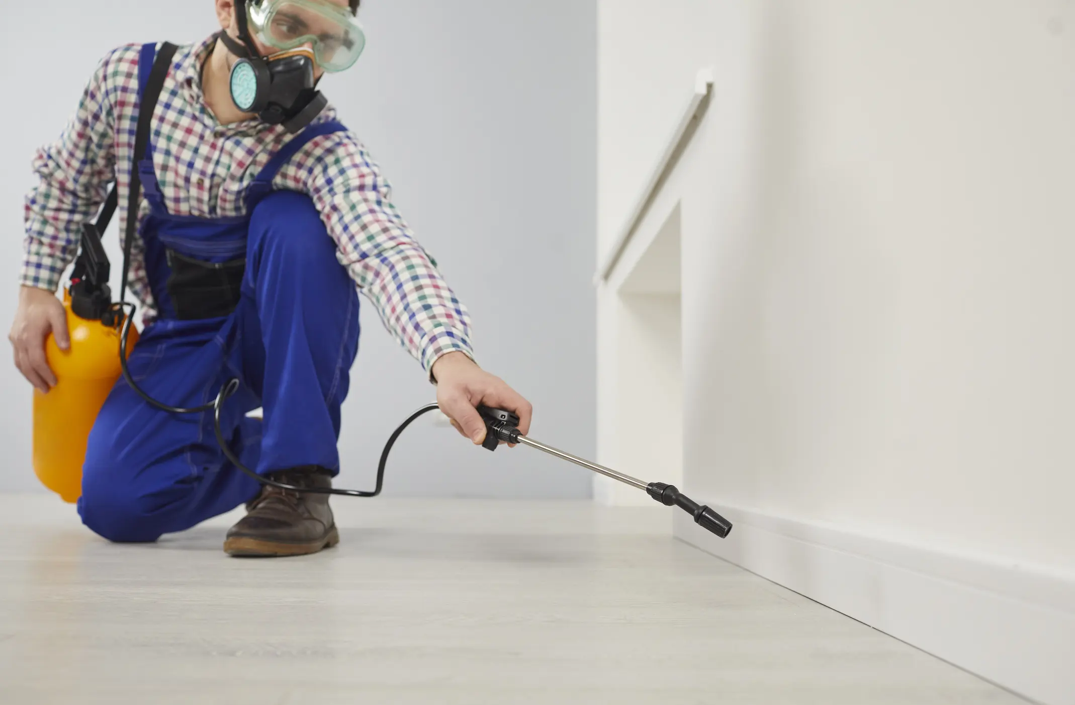 A technician wearing a plaid shirt, blue overalls, protective goggles, and a heavy-duty respirator mask kneels on a light-colored floor. He is using a professional sprayer with a long metal wand to apply treatment along the baseboard of a plain white wall. A bright yellow pressurized tank is slung over his shoulder, connected to the wand by a black coiled hose.