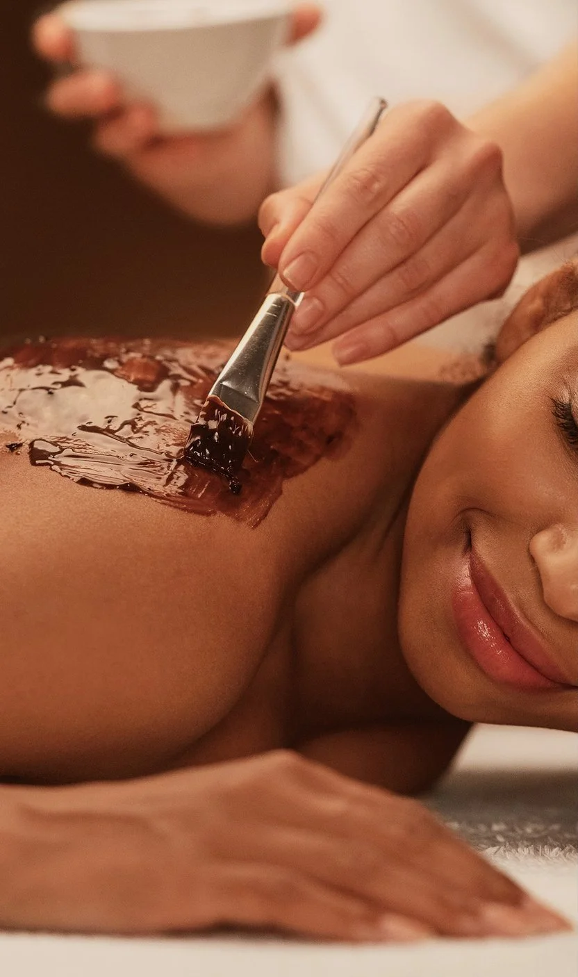 A smiling client enjoying a relaxing Herbal Blend Body Wrap as an esthetician applies the treatment to her back with a brush.