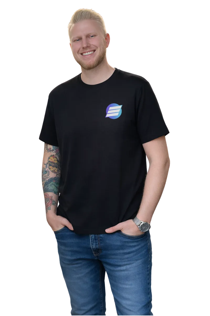 Smiling man with blond hair wearing a black t-shirt with a blue logo on the chest, blue jeans, a wristwatch, and tattoos on his left arm.