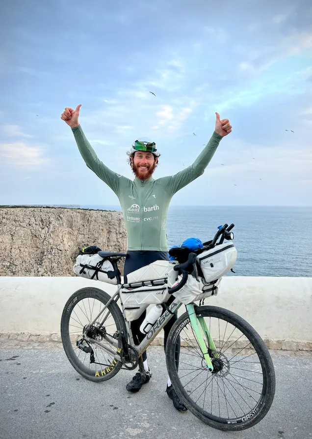 Mountainbiker hebt triumphierend die Arme, stehend auf Schotterweg vor Bergkulisse