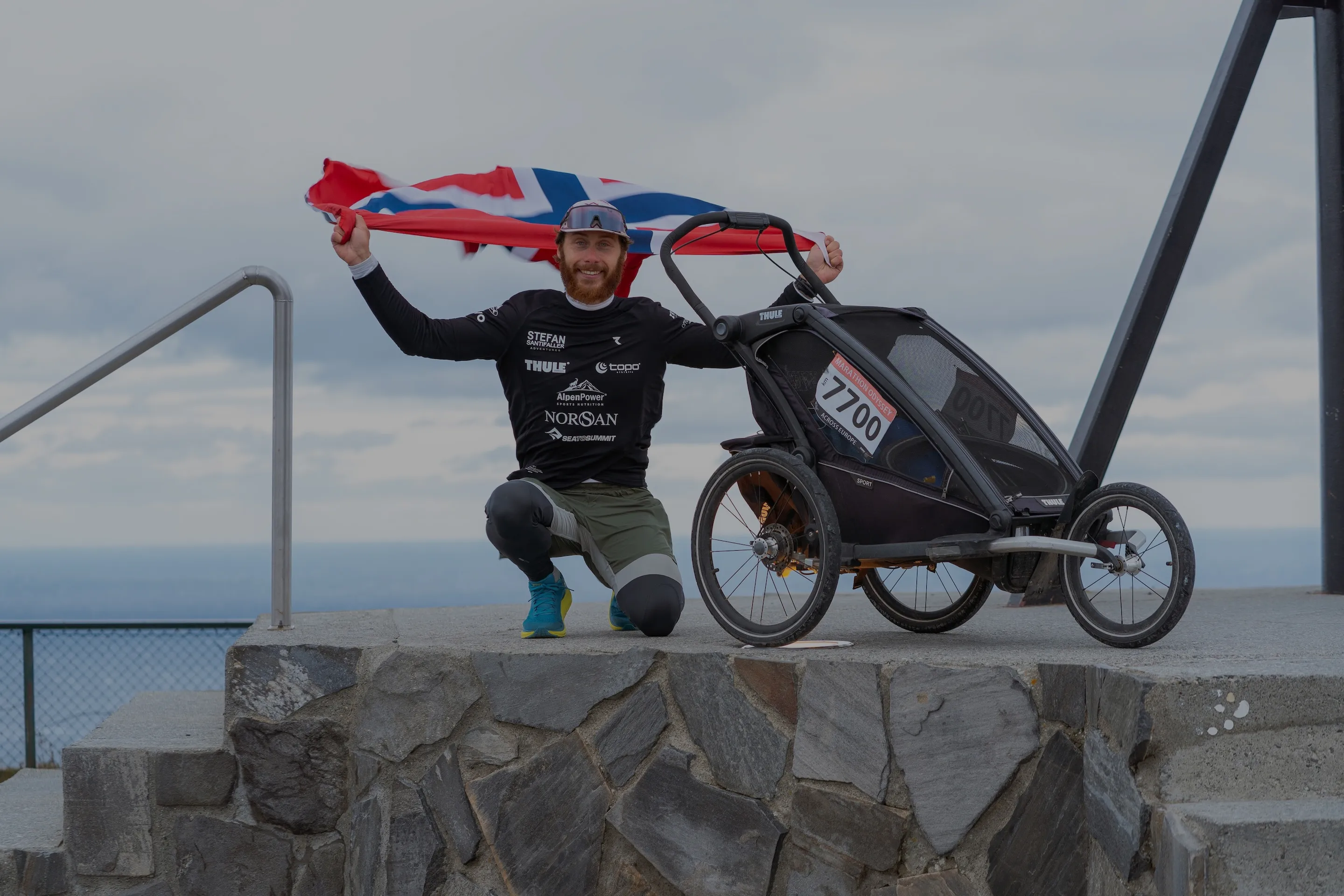 Athlet steht mit Norwegen-Flagge und Jogging-Stroller am Monument am Nordkap