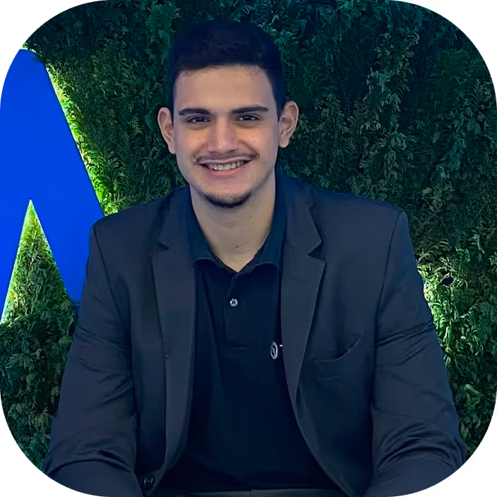 Young man with short dark hair wearing a black blazer and shirt, smiling against a green leafy background with blue lighting.