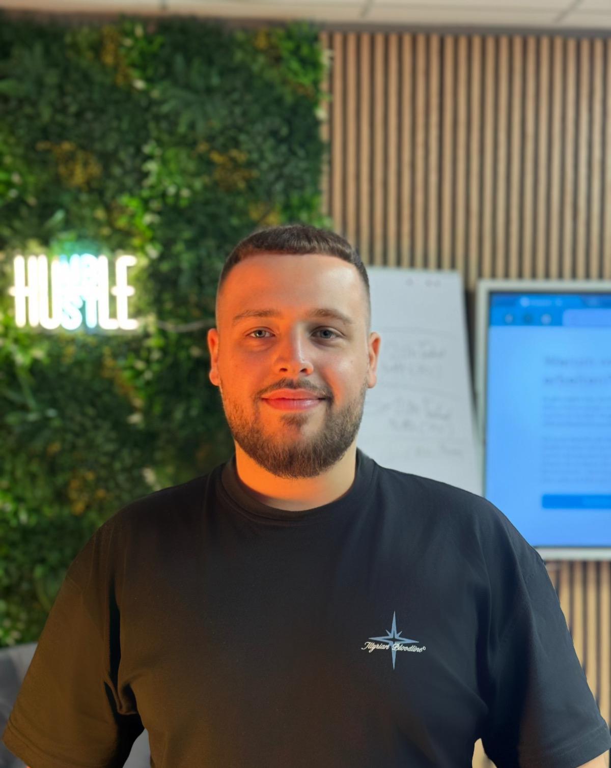 Lirigzon H., Gründer von SITEGLOW, vor Firmenwand mit Hustle-Neon-Logo