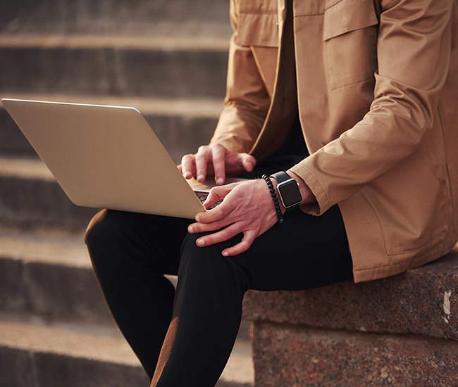 Person wearing a tan jacket and black pants sitting on stone steps using a laptop.