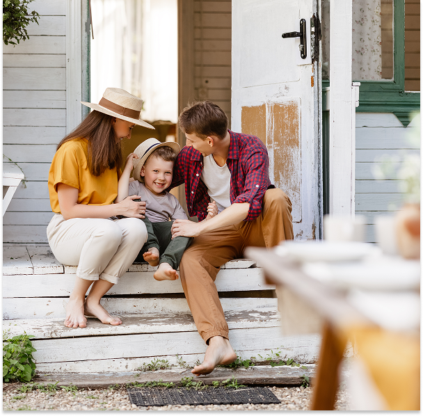 A smiling family of three sitting on wooden porch steps, with the child wearing a hat and parents engaging happily.