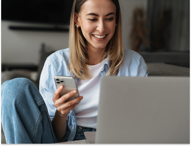 Smiling young woman sitting cross-legged using a smartphone and laptop at home.