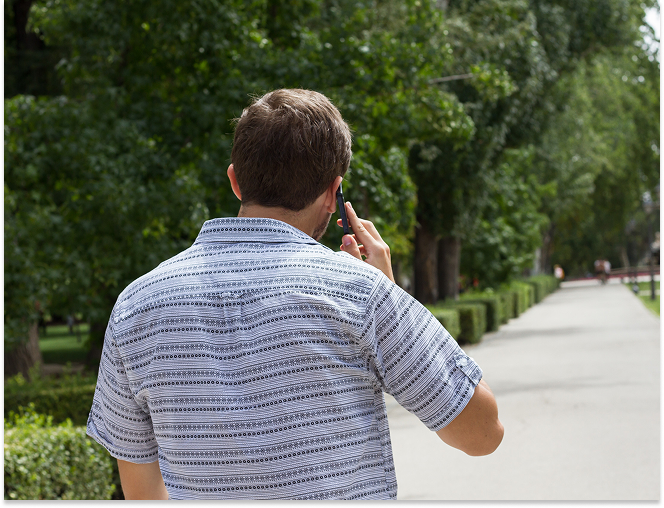 Man with short brown hair wearing a patterned short-sleeve shirt, holding a phone to his ear while walking along a tree-lined path.