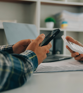 Two people sitting at a desk, each holding a smartphone over documents with a laptop and shelves in the background.