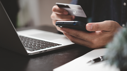 Person holding a credit card and a smartphone near a laptop on a desk.