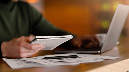 Person holding a notepad with laptop and papers on a desk in a blurred background.
